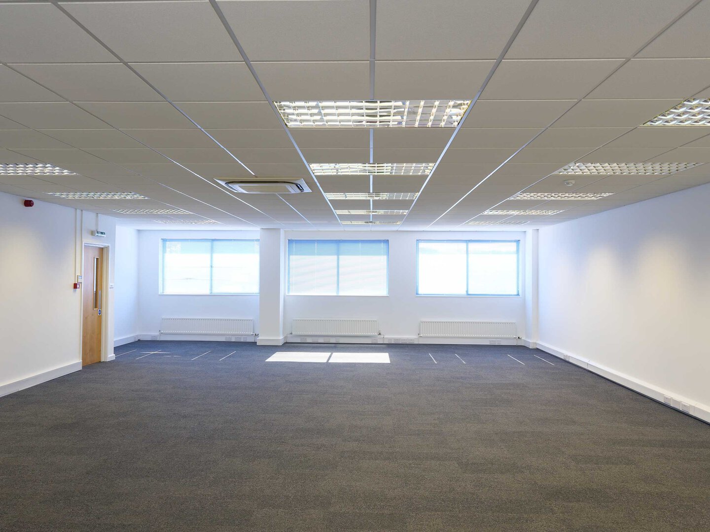 Empty modern office space with gray carpet, white walls, and large windows providing natural light.