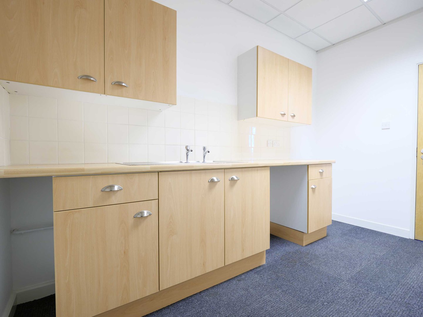 Modern office kitchenette with light wood cabinets, white countertop, and double sink.